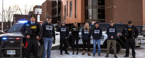 Police foundations students and cadets standing with patrol vehicles outside a campus building in winter.