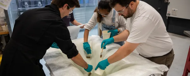 Students wearing safety gear apply resin to a fiberglass mold during a hands-on composites lab