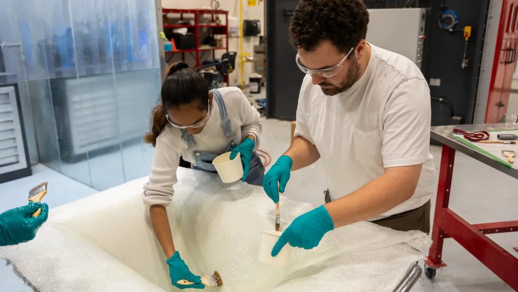 Students wearing safety gear apply resin to fiberglass fabric in a hands-on composites manufacturing lab.