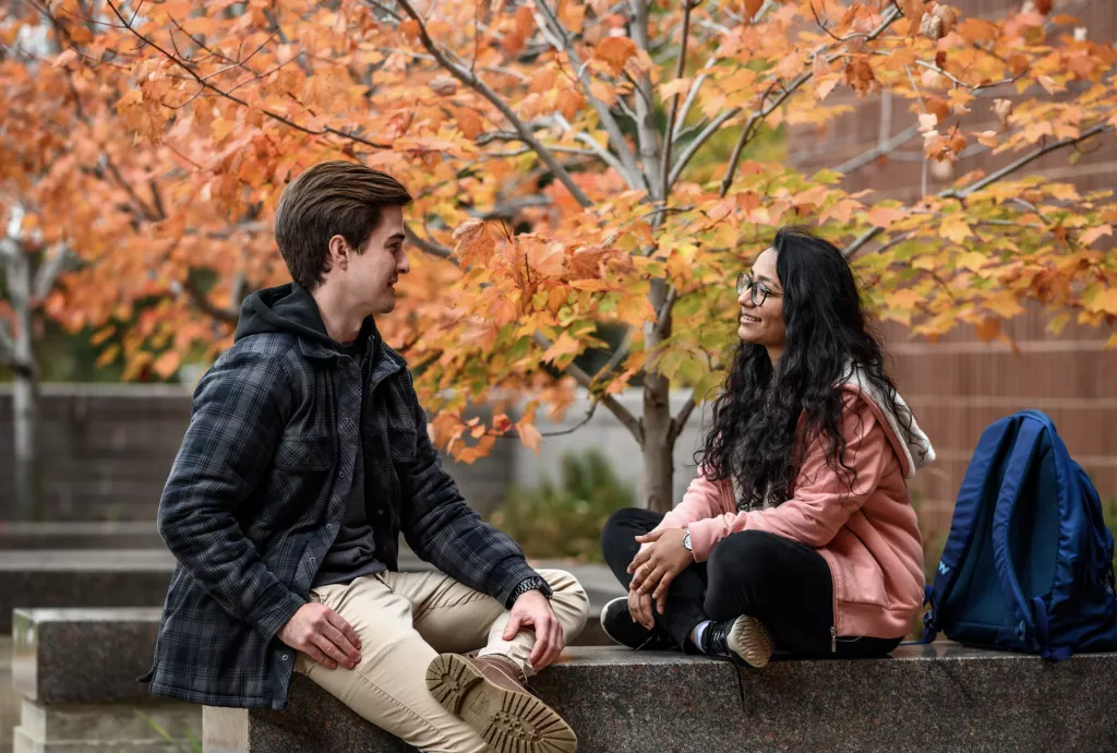 Two students sitting safely in front of the SAFE campus at Confederation College with a fall, brown leafed tree in the background.