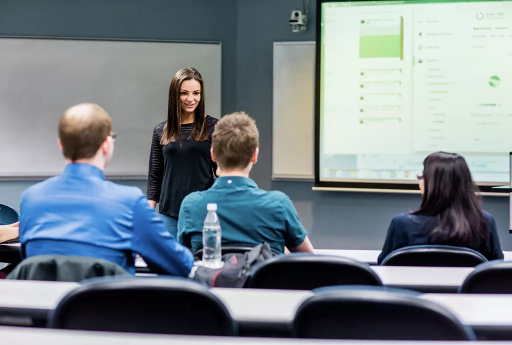 A lecturer giving a presentation to a group of attentive students, with a chart visible in the background.