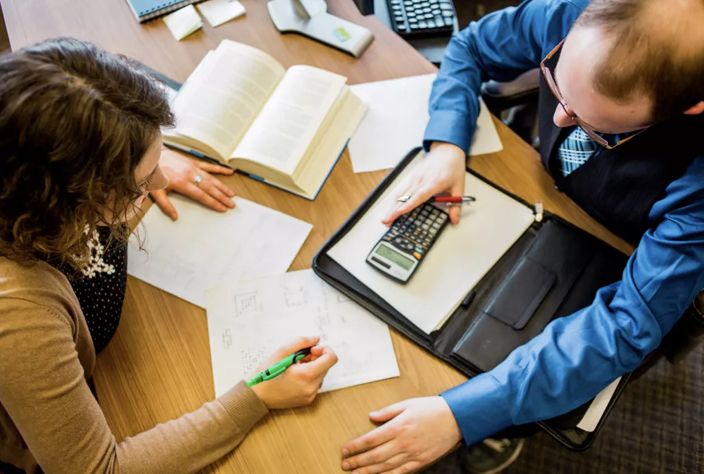 Two people, a man and a woman, sitting at a table with calculators and notebooks, engaged in accounting and finance activities.