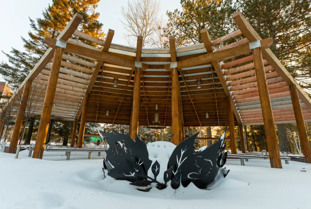 Circular wooden outdoor pavilion with tiered benches, surrounded by trees and snow on campus in winter