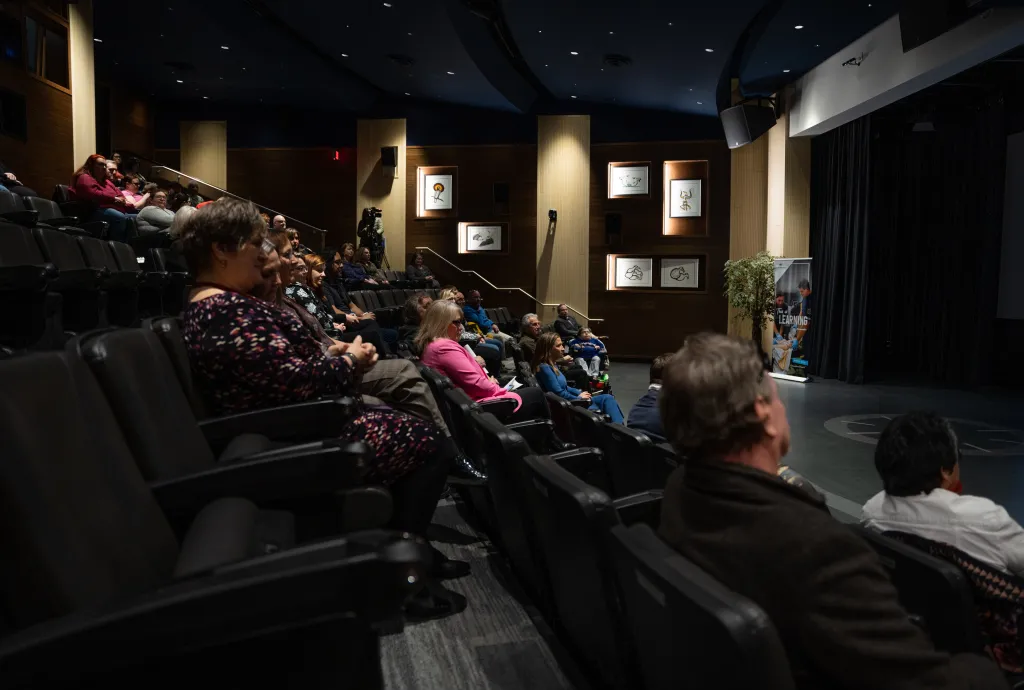 People seated in an auditorium at Confederation College, attentively watching a presentation. The room is dimly lit, with artwork on the walls and a banner visible near the stage.