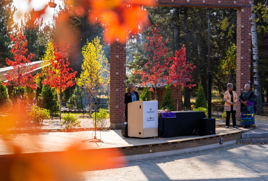 A woman speaks at a podium on an outdoor campus stage while two other people stand nearby. Autumn trees with red and yellow leaves frame the scene, and Confederation College branding is visible on the podium.