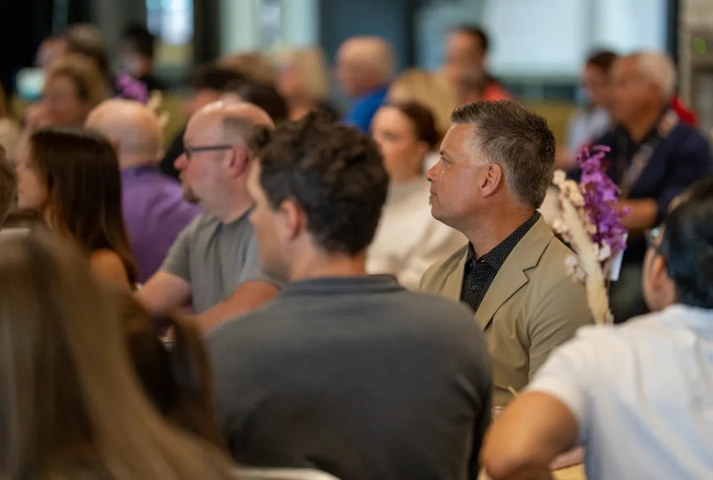 A group of faculty and staff seated indoors, attentively listening during a college event. They are dressed in casual to business attire, with decorative flowers visible on the tables.