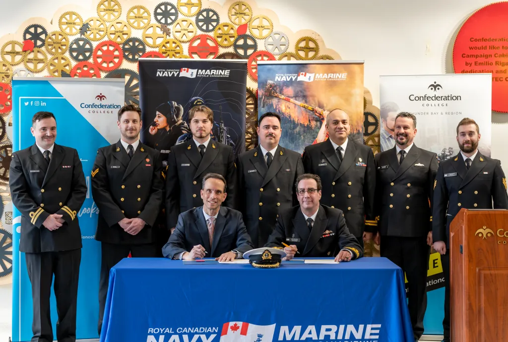 Royal Canadian Navy personnel and officials sign agreement at Confederation College with banners in background