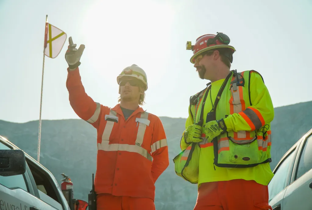 Two workers in safety gear at an industrial site, one pointing while discussing work beside trucks.