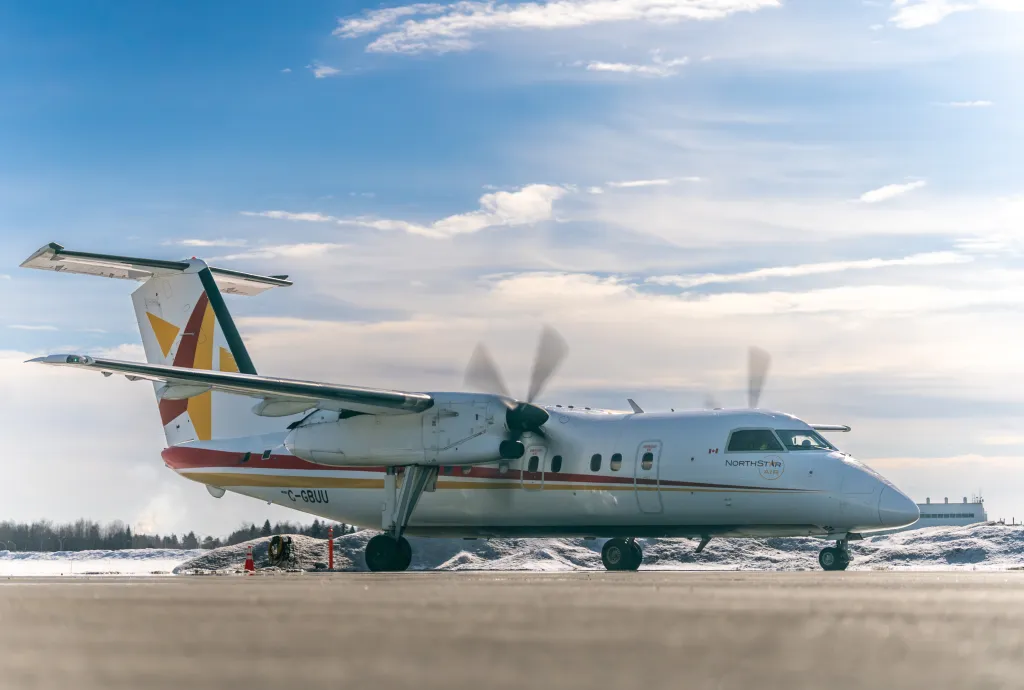 A white North Star Air turboprop airplane sits on a snowy runway under a bright sky. The aircraft is shown in profile with its propellers blurred from motion and colored stripes on the tail and fuselage.