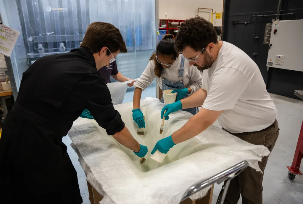 Students wearing safety gear apply resin to a fiberglass mold during a hands-on composites lab