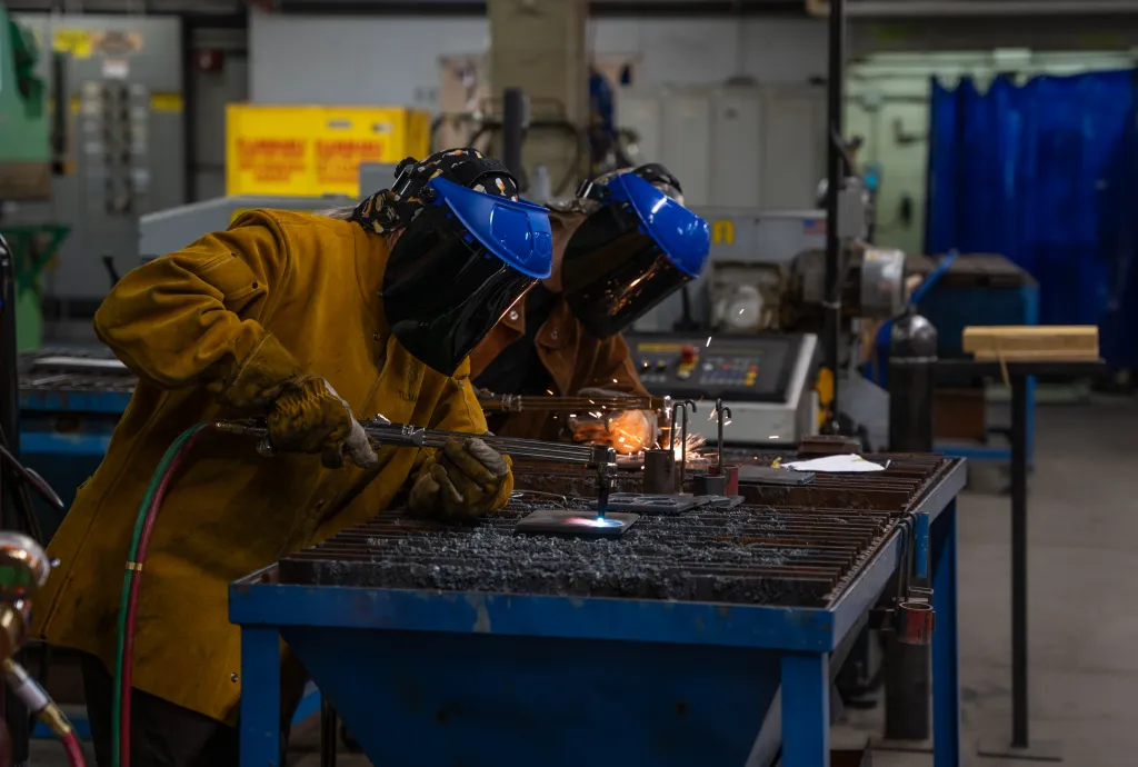 Two students wearing welding helmets and heavy protective jackets work at a metal welding station in a trades lab. One student is actively welding as sparks fly across the work surface, with industrial equipment and blue safety curtains visible in the background.